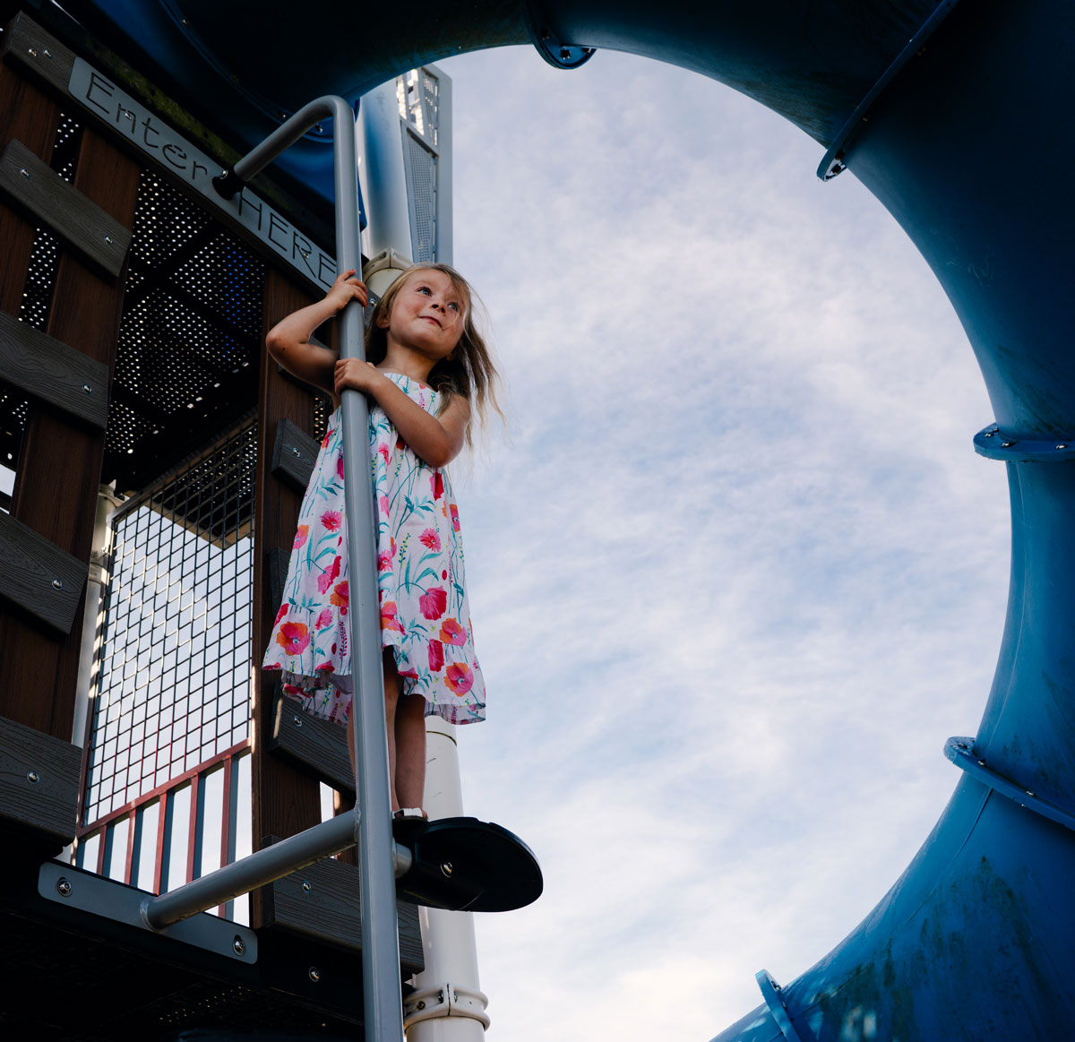 Izzy Newtown, in a flowery dress, standing on a playground pole with the sky visible prominently through the image behind her