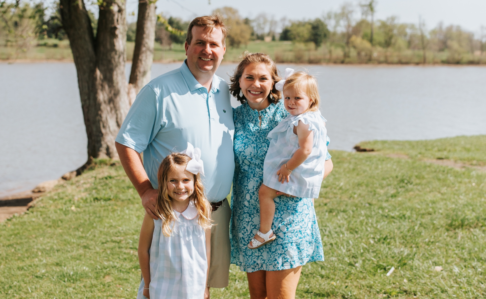 A photo of the Parrish family smiling as they pose together for the camera in front of a lake at the park.