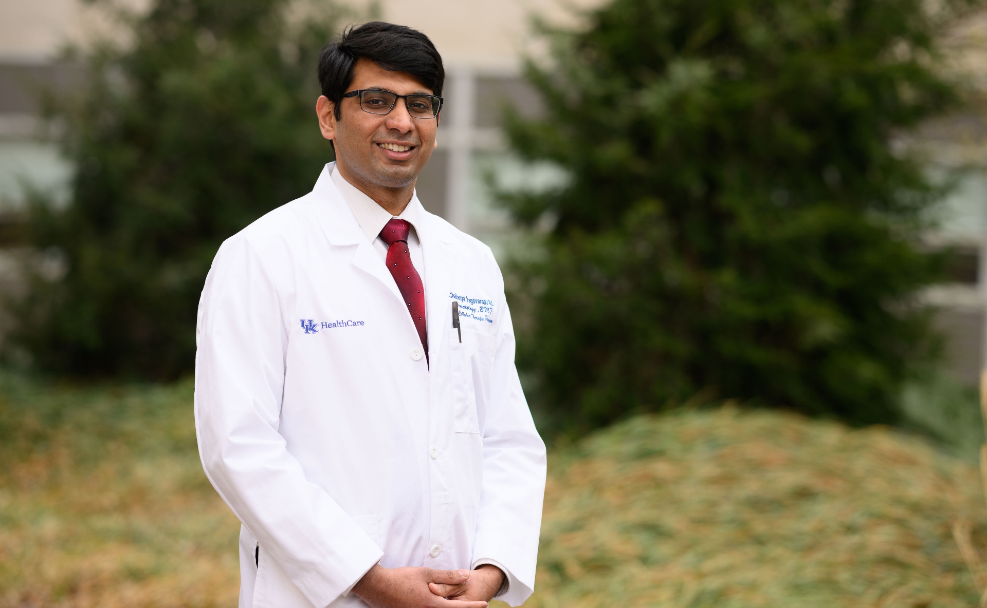 A photo of Dr. Chaitanya Iragavarapu smiling as he stands outside in front of a building on the UK HealthCare campus. He is a South Asian man with short black hair. He is wearing a white lab coat over a white button-up shirt with a red tie.
