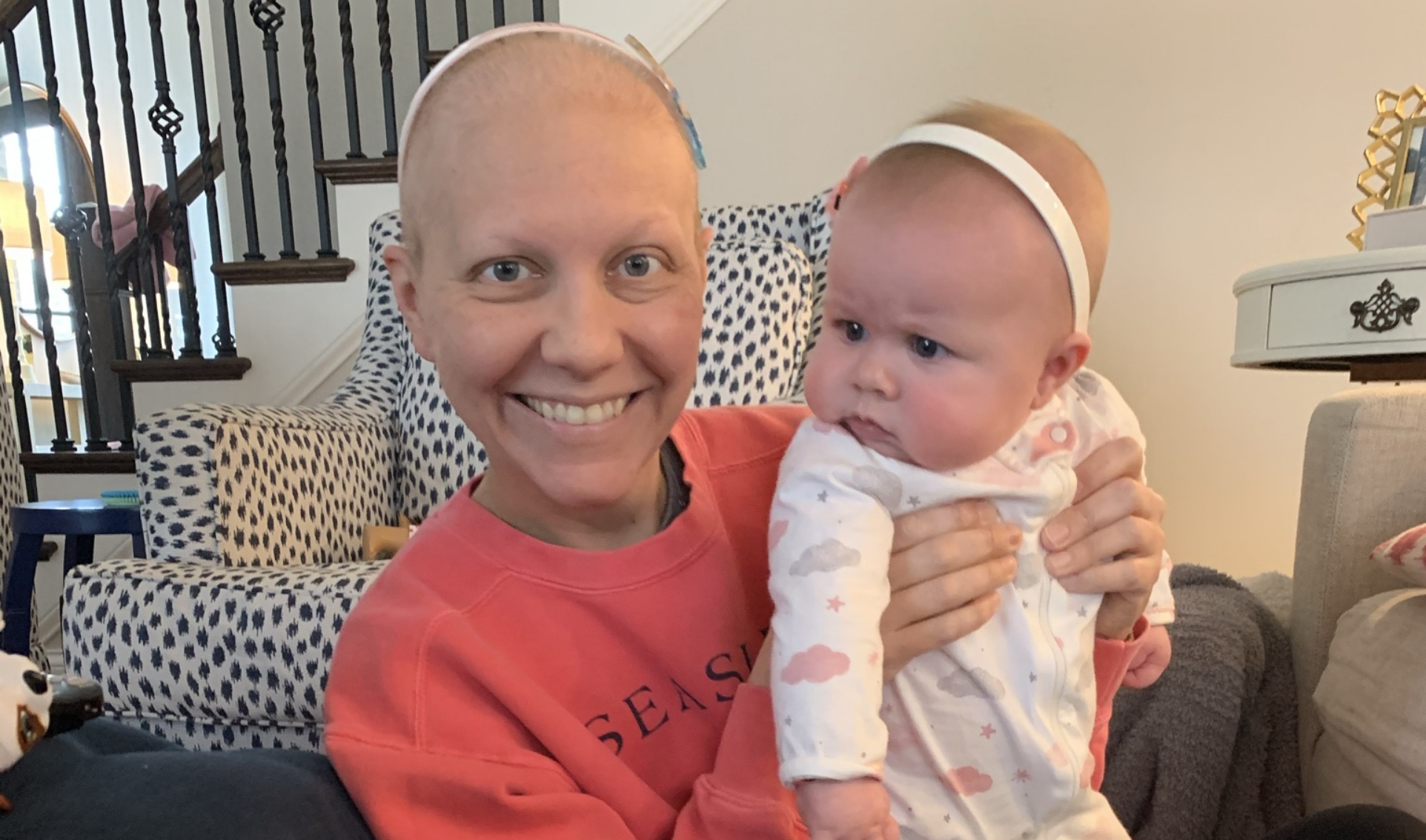A close-up photo of a bald Anne Sydney smiling while holding up baby Eloise. They are wearing matching white headbands.