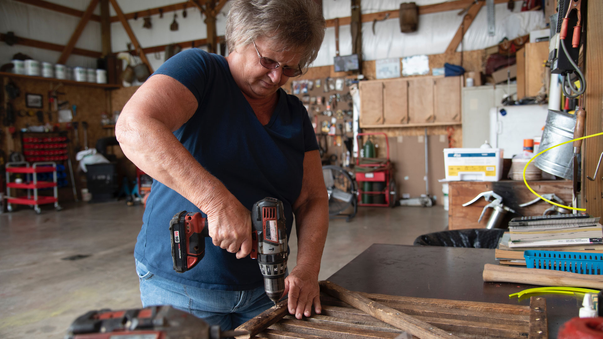 A close-up photo of Charlotte, wearing a navy blue shirt and jeans, using a power drill on a piece of wood in the barn on her farm.