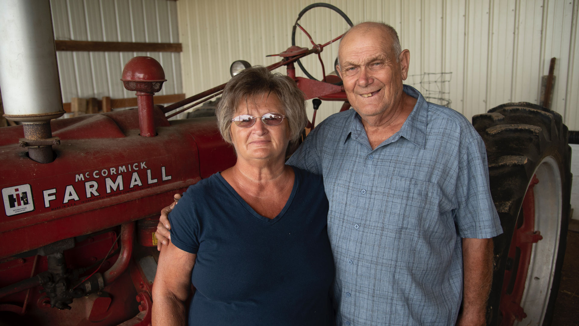 Charlotte and her husband, an older man in a blue button-up shirt, posing for a photo in front of an older red tractor.
