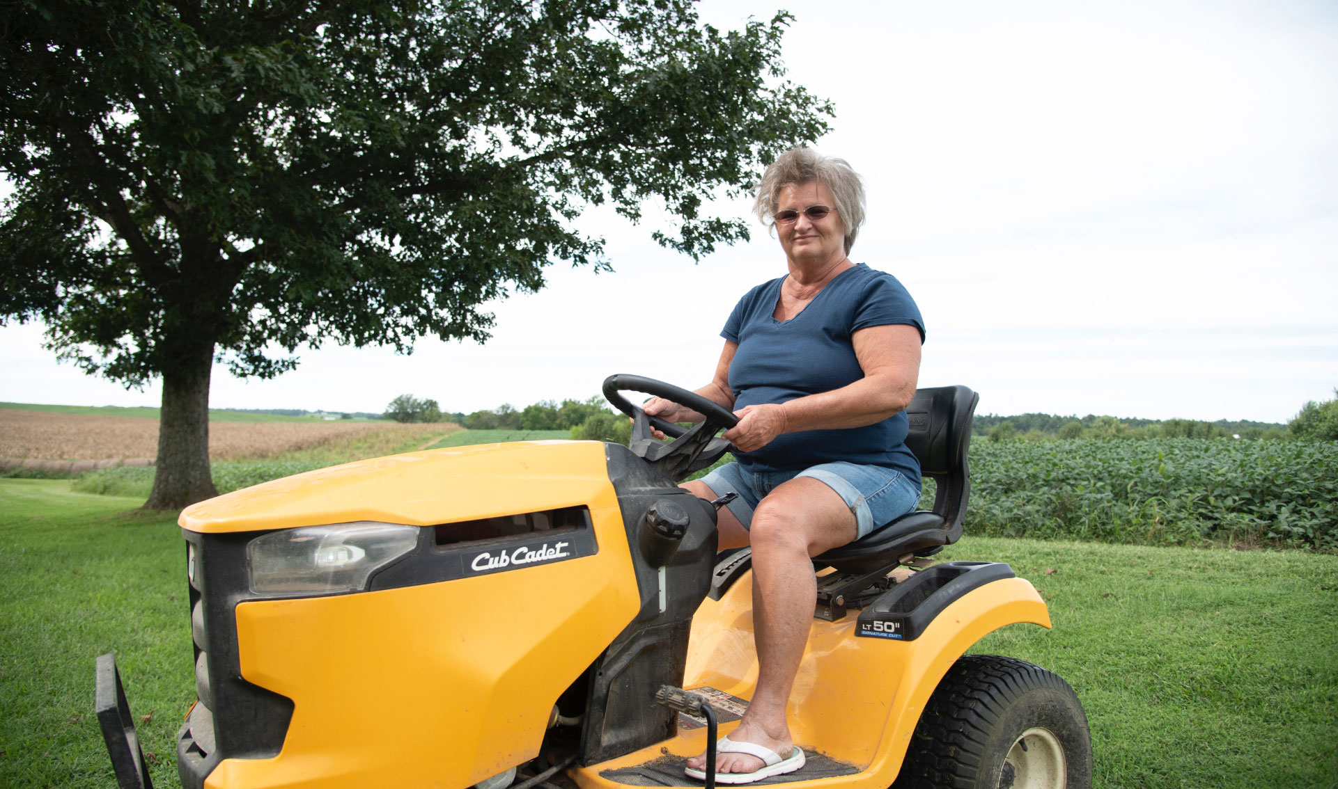 A photo of Charlotte smiling at the camera as she drives a yellow riding lawn mower.