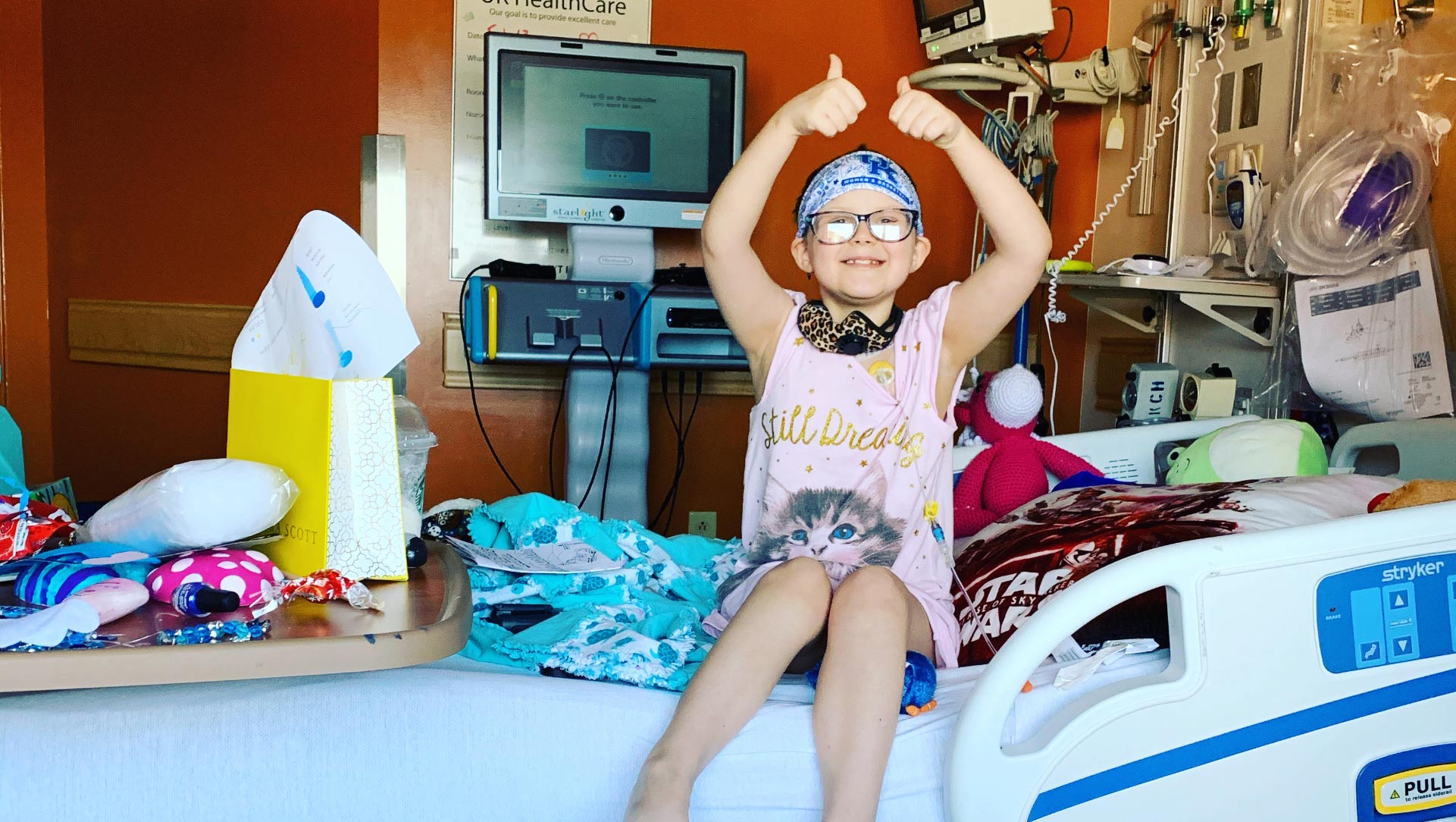 A photo of Ellie in her hospital room where she is sitting upright on the bed with both arms in the air giving two thumbs ups. Her room is full of colorful blankets, stuffed animals, and a yellow gift bag on her bed.