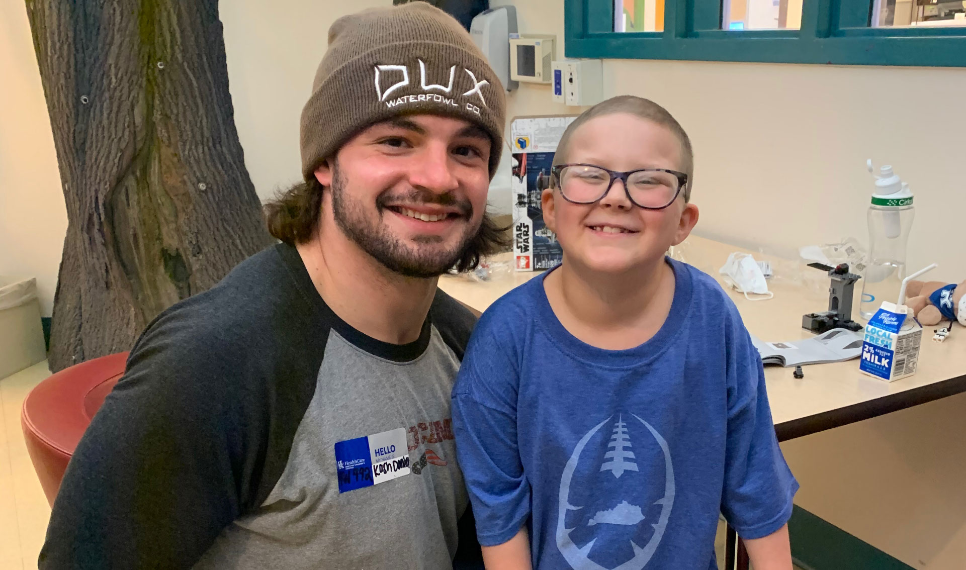 Ellie and Kash Daniel, a young man with an athletic build, brown hair, and a beard wearing a brown beanie and a gray and black t-shirt, sit together and smile for a picture.
