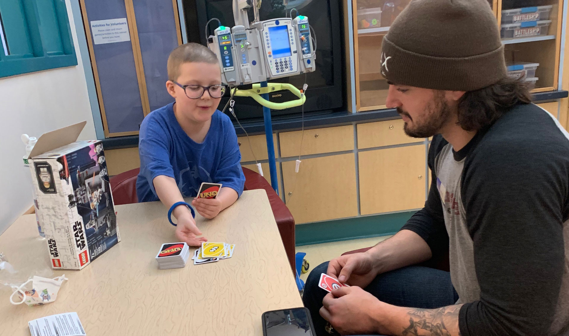 Ellie and Kash sit together at a table in the hospital and play Uno together.