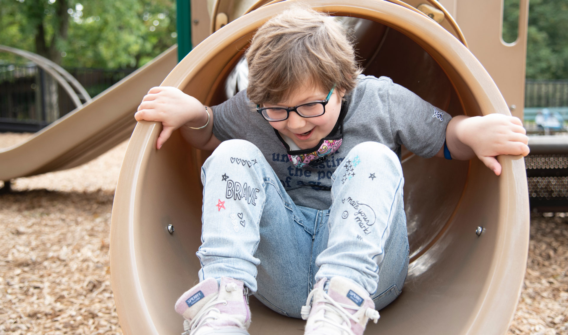 A photo of Ellie at the bottom of a tunnel slide with her hands grabbing the outside of the slide while she enthusiastically smiles. She is wearing a gray t-shirt, jeans, and light pink sneakers.