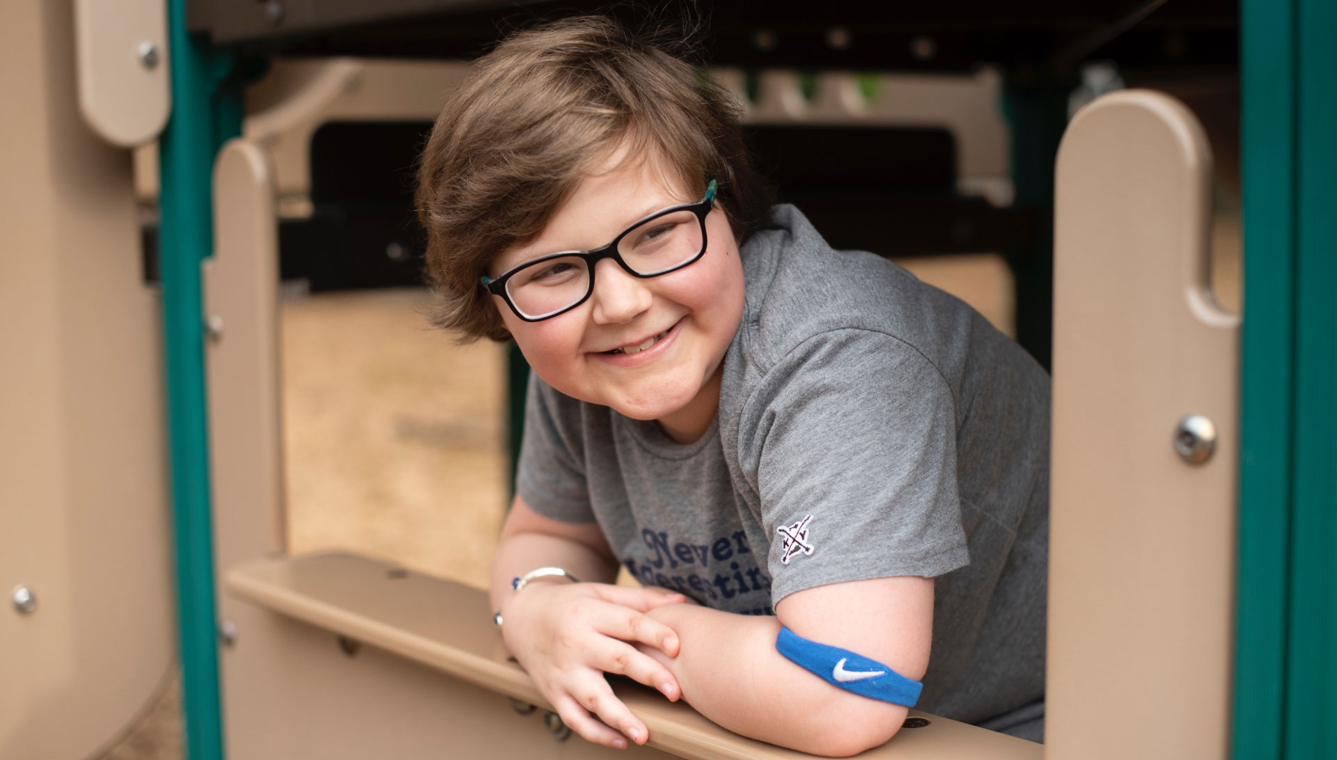 A portrait of Ellie Hurley, a young girl with short brown hair and glasses, wearing a gray t-shirt, posing on a playground. She is smiling while looking away from the camera.