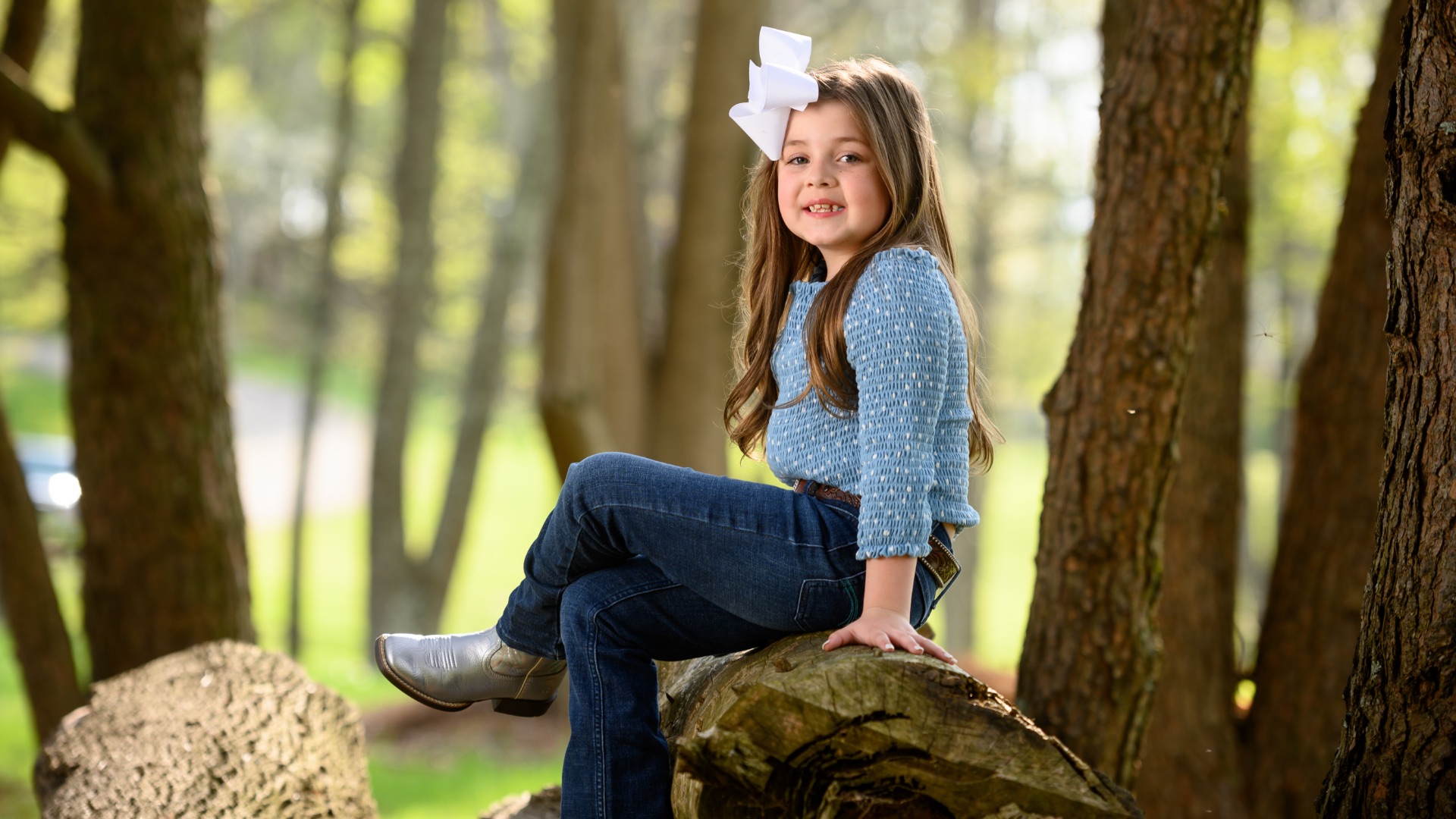 Henley Thurman, a 6-year-old white girl with brown hair, smiles as she sits on a broken tree trunk with her legs crossed. She is wearing grey boots, navy jeans, light blue top and a big white bow in her hair.