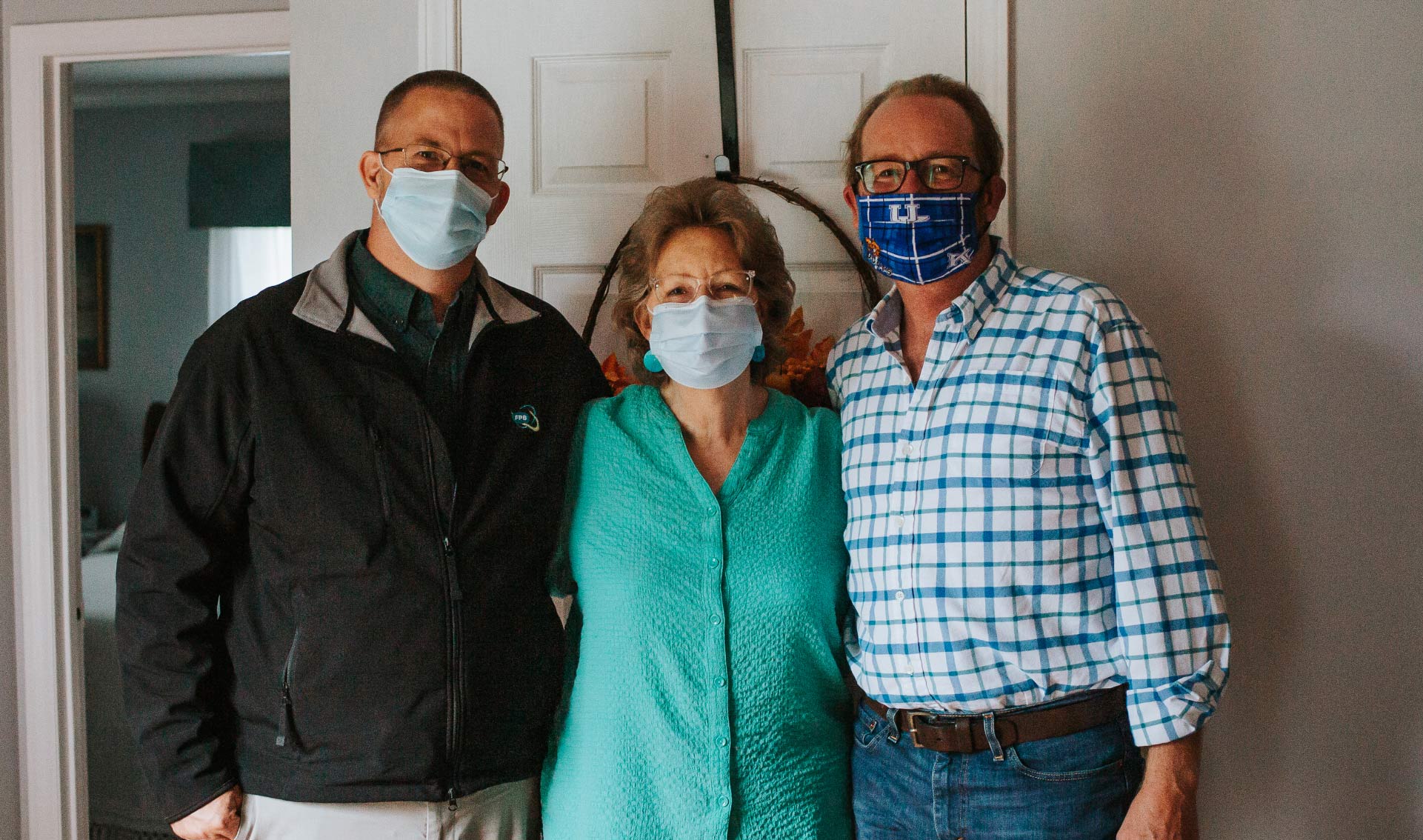 Mae standing closely between her sons. On her left is Kenneth, a middle-aged white man wearing a black jacket, glasses and a facemask. On her right is Scotty, in a white and blue checkered shirt, black framed glasses and a facemask.