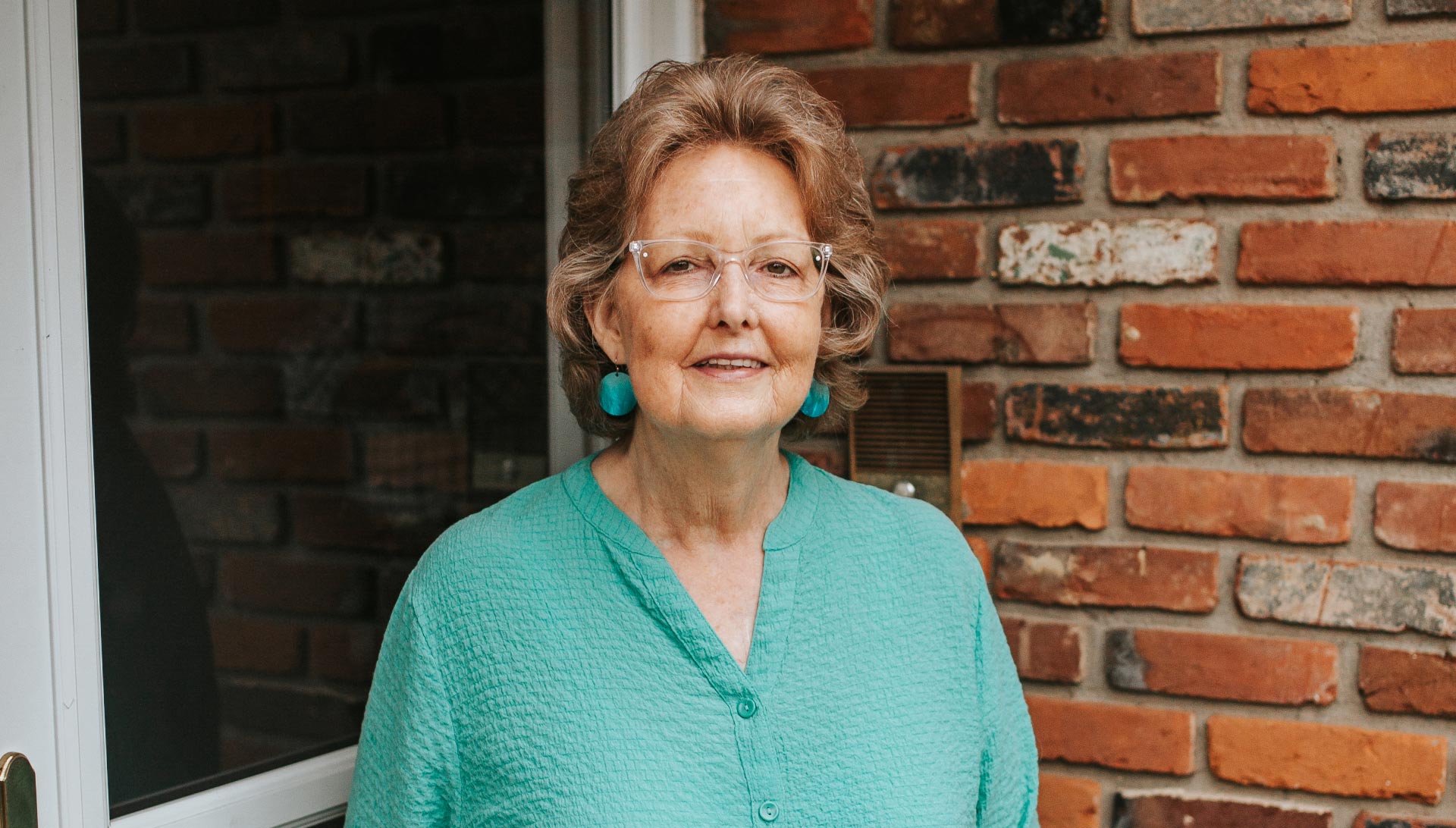 Mae Lewis, an elderly white woman with brown and grey hair, stands in an outside setting area. She is wearing clear framed glasses and a turquoise shirt with matching earrings.