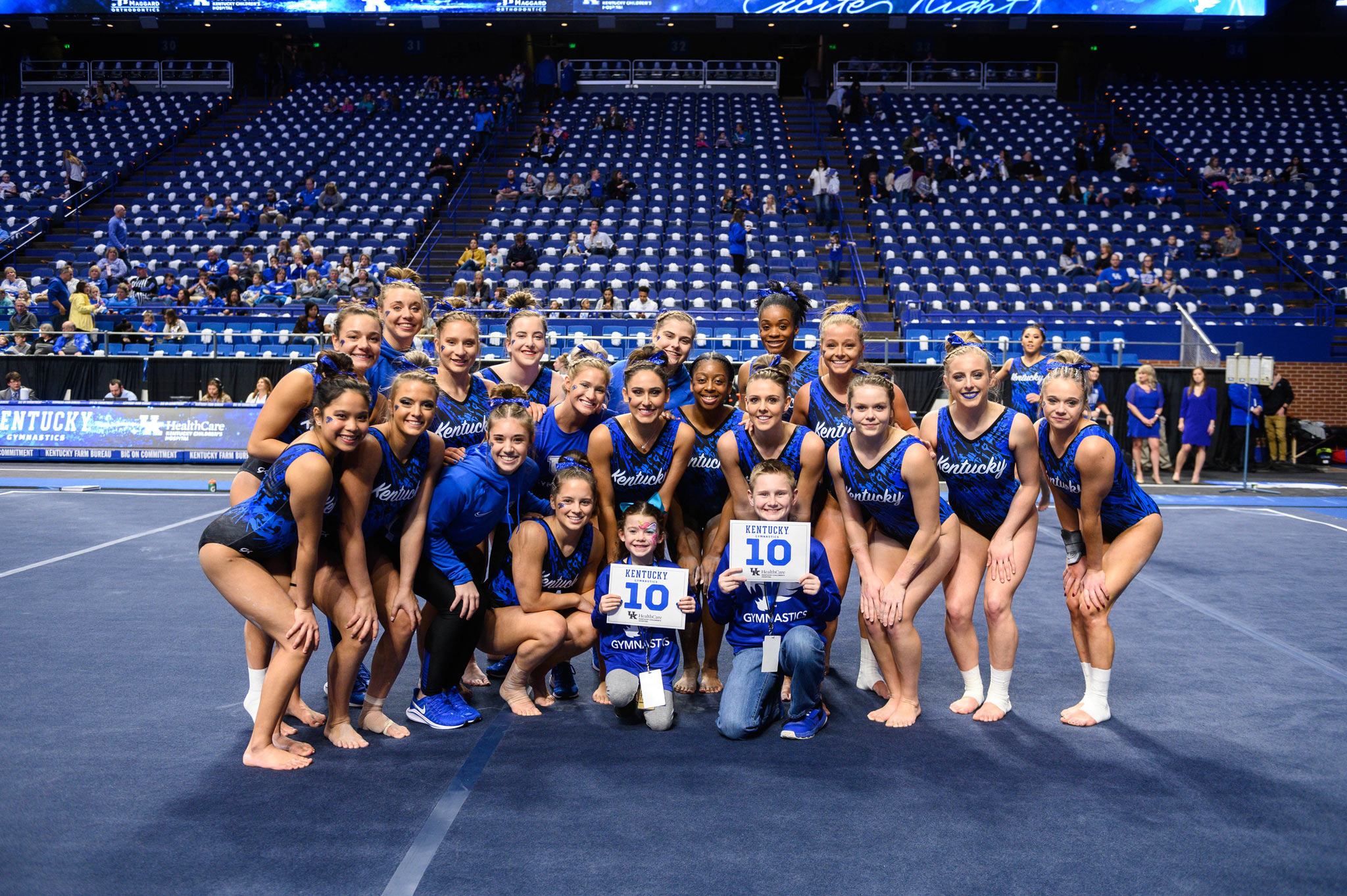 Izzy and Dalton hold signs with 10's on them and kneel in front of a group of UK gymnasts.
