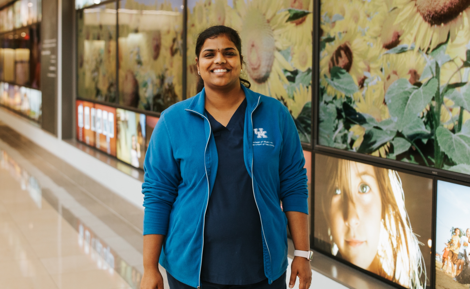A photo of Dr. Rani Vasireddy smiling while standing in front of a wall decal with sunflowers on it. She is a South Asian woman with long black hair that is tied back in a ponytail. She is wearing a blue UK HealthCare-branded jacket over a pair of dark blue scrubs.