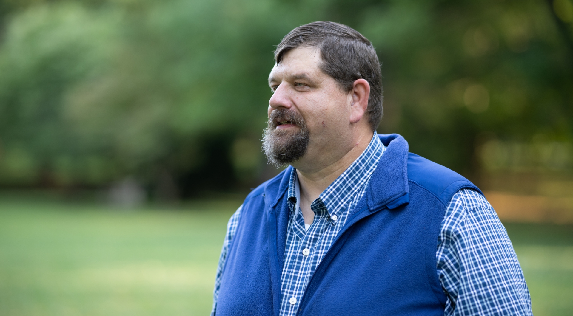 A close-up photo of Ronald Mullins looking off into the distance while standing outside. He is an older white man with short gray hair and a gray goatee. He is wearing a blue vest over a blue long-sleeve checkered shirt.