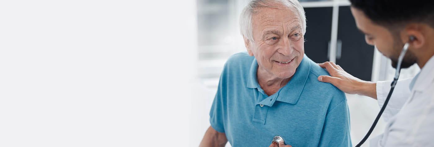 An older man wearing a light blue polo shirt sits on an exam table as a younger, male doctor uses a stethoscope to listen to his chest.