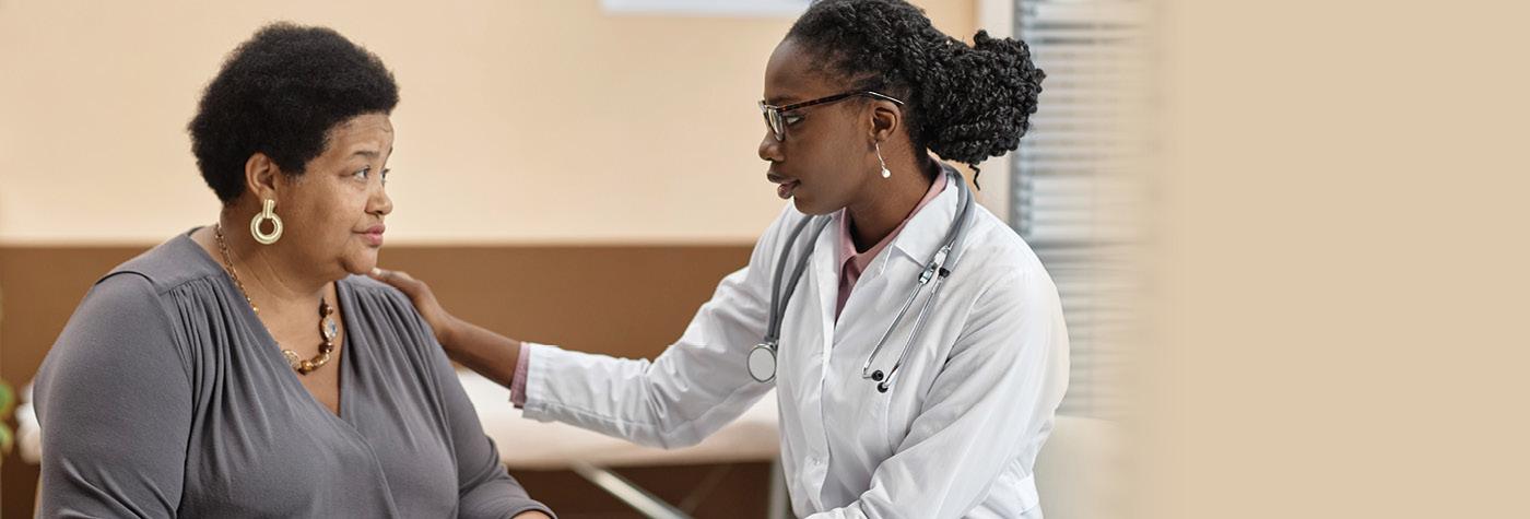 A middle-aged African-American female patient wearing a grey blouse has a conversation with her provider, a younger African American woman wearing a white coat with a stethoscope draped over her shoulders.