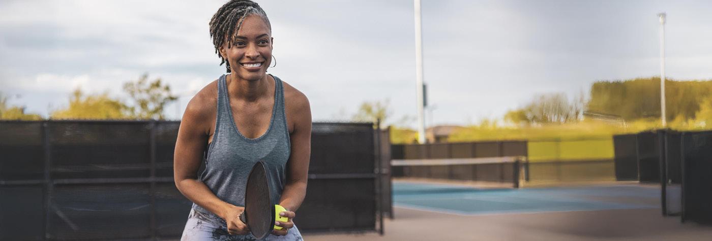 A smiling woman wearing a gray tank top holds a tennis racket.