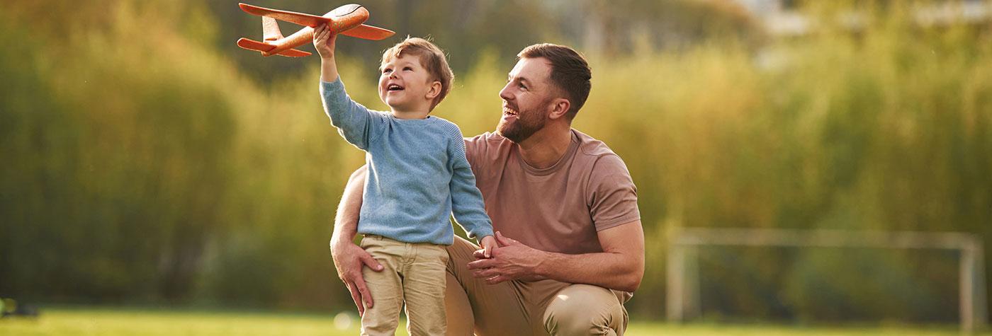 A father and his young son laugh together as they play outdoors with a toy airplane.