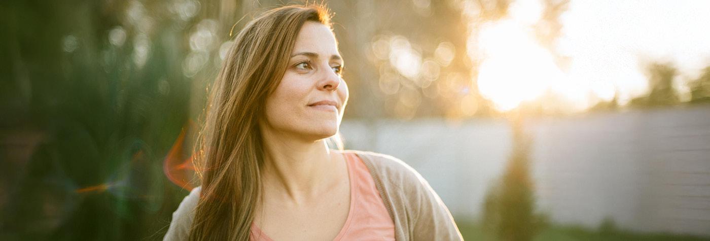 A woman wearing a pink shirt and tan cardigan gazes to her left as she stands outside. A tree and stockade fence can be seen in the background of the image as the sun begins to set overhead.