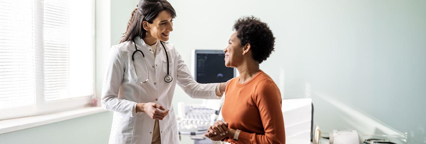 An African-American woman wearing an orange, long-sleeved shirt smiles as she speaks with her doctor, a woman wearing a white coat with a stethoscope draped around her neck. They are in an exam room. 