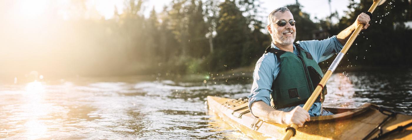 A man with graying hair and a beard paddles a kayak down a river while wearing sunglasses, a green life vest, and a light blue chambray shirt.