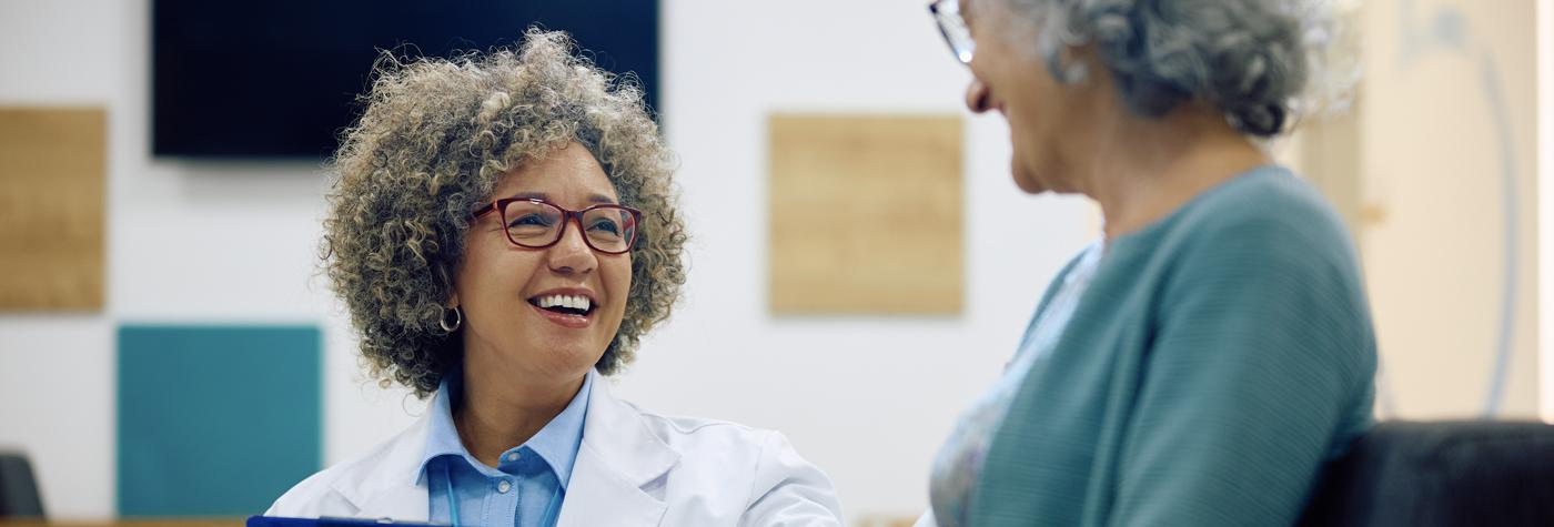 A woman and her doctor (also a woman), smile as they speak with one another.