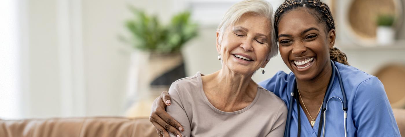 An older woman smiles while sitting next to her healthcare provider, a black woman wearing light blue scrubs and a stethoscope draped around her neck. 