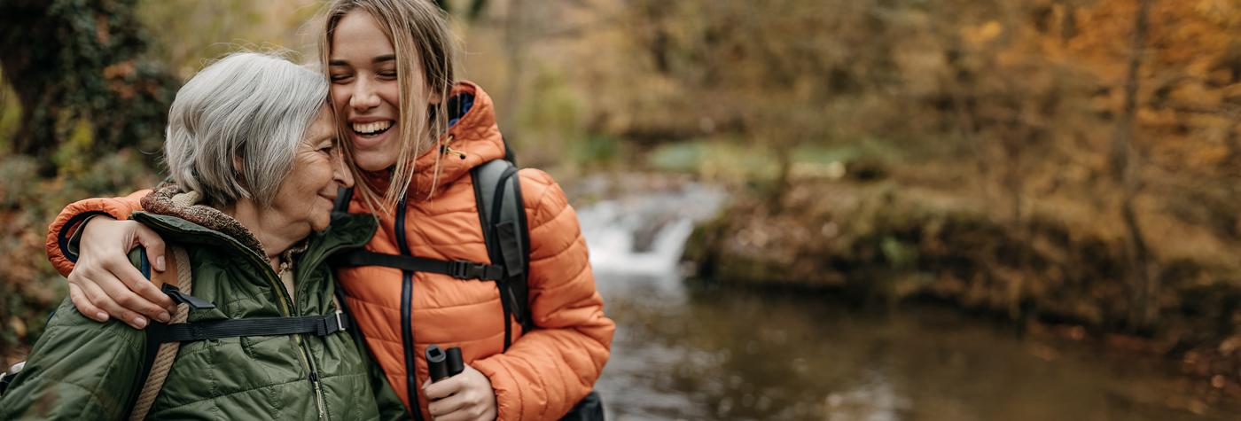 An older mother and her adult daughter embrace as they stand by a river in their hiking gear. The vivid orange and yellow leaves in the background show that the photo was taken in the autumn.