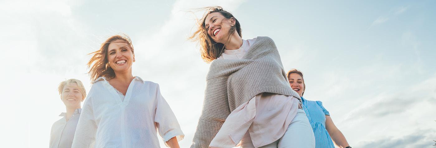 Four smiling women walk in a group with a partly cloudy sky in the background.