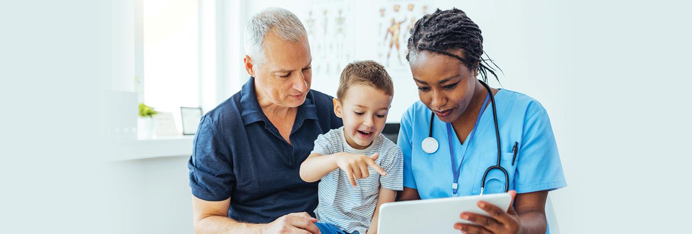 A man holds his young son on his lap while they look at a tablet computer being held by the child's doctor, an African American woman wearing light blue scrubs with a stethoscope draped around her neck. 