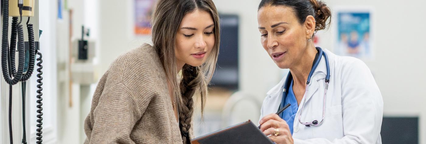 A young woman speaks to her doctor, a woman wearing a white coat with a stethoscope draped around her neck.