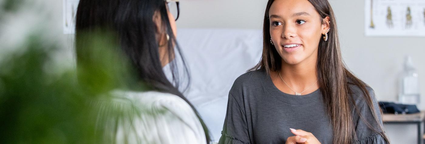 A teenage girl with long, dark hair speaks to her doctor. 