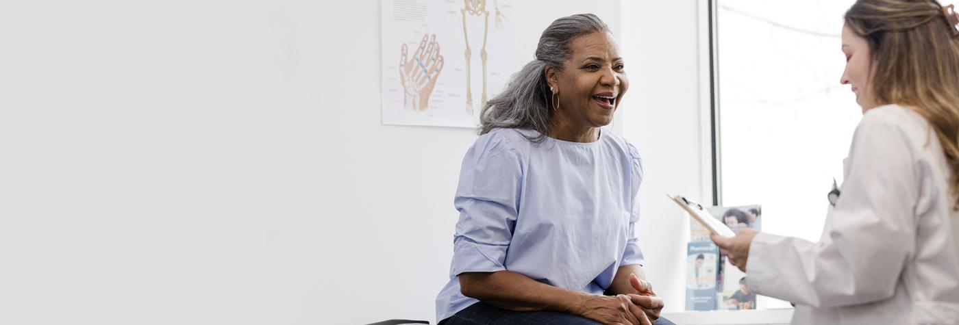A middle-aged African-American woman sits on an exam table and talks to her doctor, a woman with long brown hair wearing a white coat.