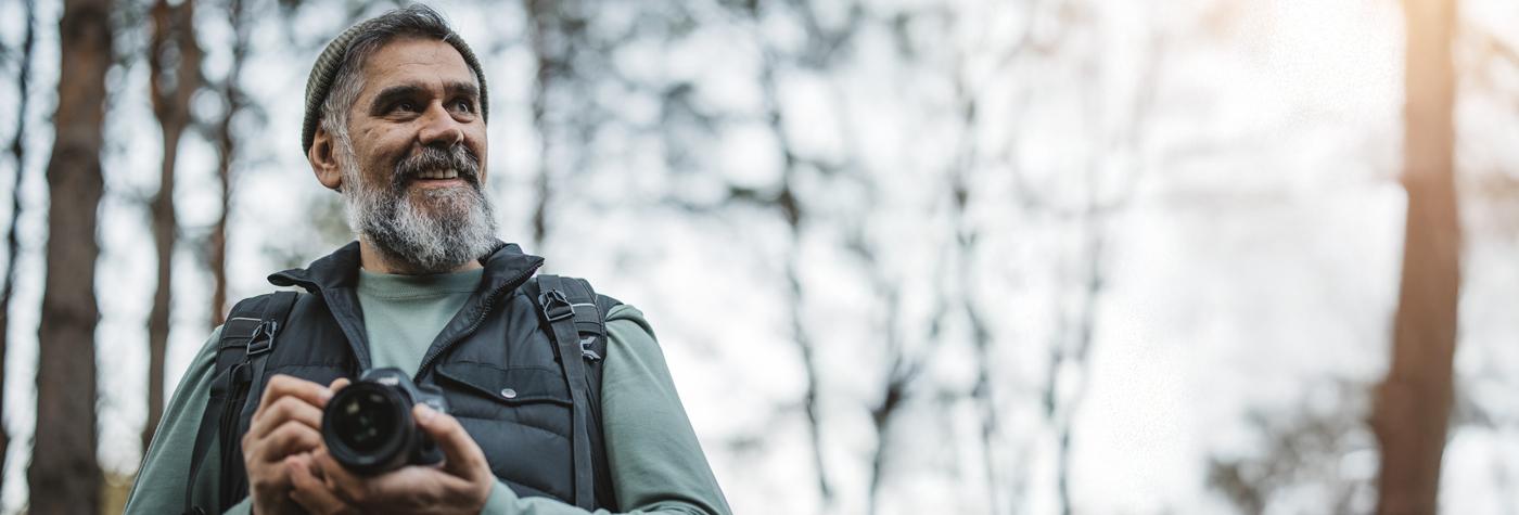 A man with a graying beard stands in the woods holding a camera in his hands.