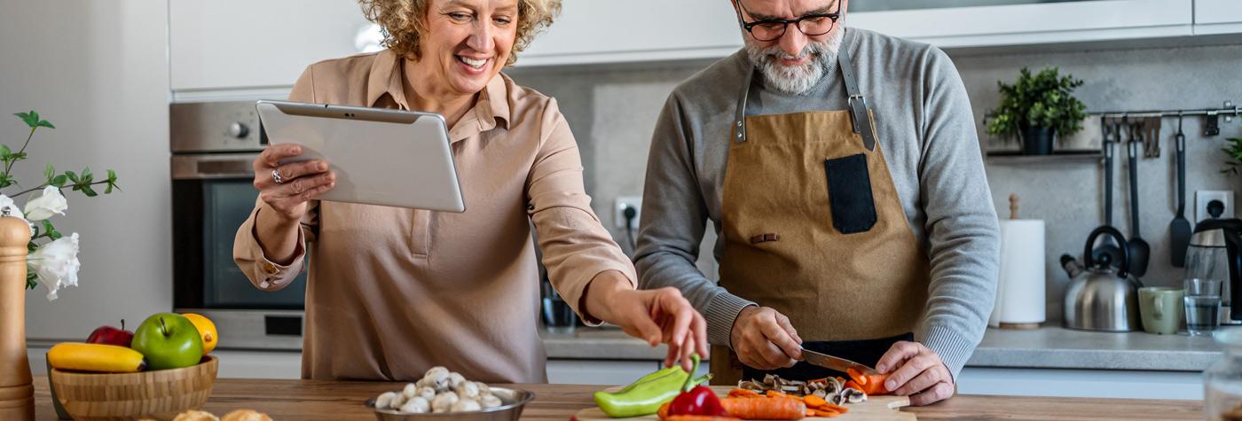 A woman holding a tablet stands next to a man who is wearing an apron and chopping vegetables on a kitchen counter.