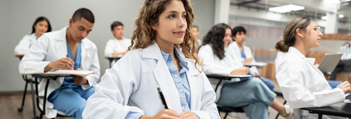 Several people in white lab coats sit in rows of desks in a classroom setting, writing notes while facing forward.