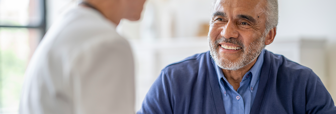 Close-up of a person with short gray hair and a gray beard, wearing a blue shirt and cardigan, seated indoors and facing another person in a light-colored top who is out of focus.