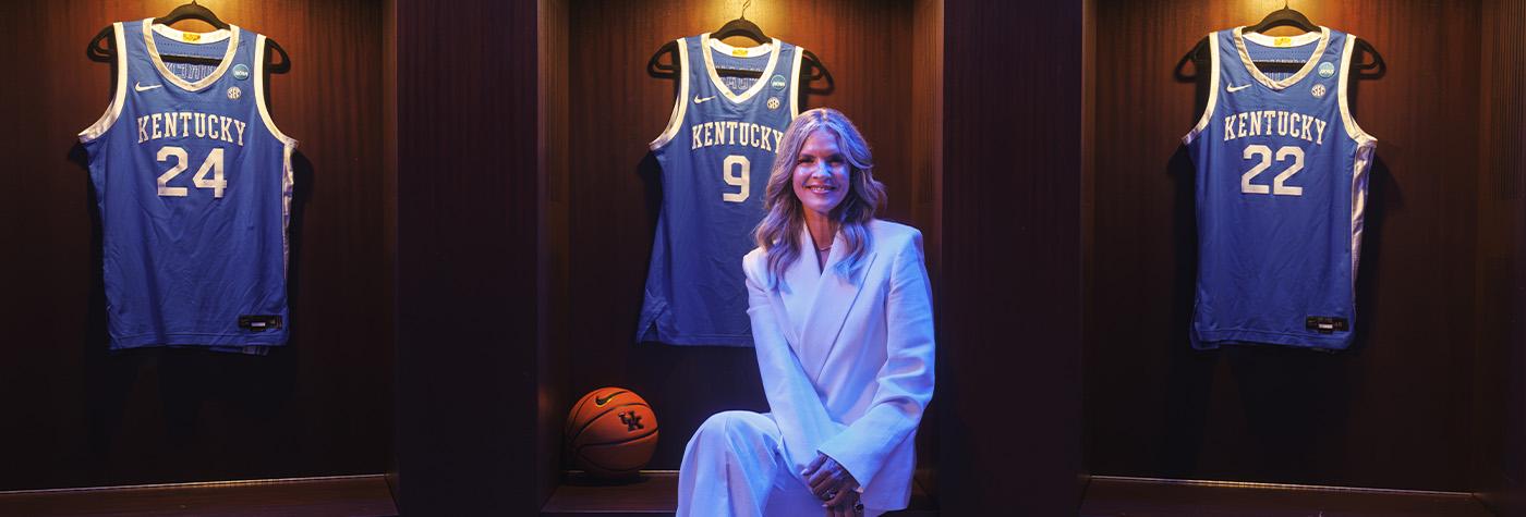 Lee Anne Pope sits in front of several Kentucky men's basketball jerseys in the UK Men's Lockerroom