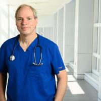 Dr. Michael Anstead, a middle-aged white man with grey hair, stands in the hallway of UK HealthCare wearing blue scrubs with a stethoscope around his neck.