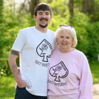 Cameron and his grandmother, an older white woman with white hair, stand and smile side by side. She is wearing a pink long sleeved ‘Team Cam’ shirt with her spectacles on her head.