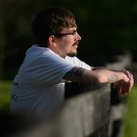 Cameron stands turned to his side, with his hands on the border fence of the park as he stares into the horizon.