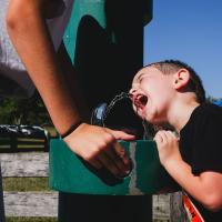 Granger Smith, right of frame, drinks from a water fountain as his mom, visible partially in the frame to the left, holds the water fountain activator button