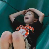 Granger Smith slides down the end of a playground slide, excited with his hands over his head.