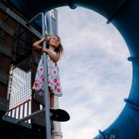Izzy Newtown, in a flowery dress, standing on a playground pole with the sky visible prominently through the image behind her