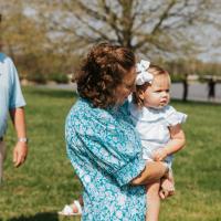 A photo of Anne Sydney holding her youngest child, Eloise, on her hip. Her husband, Jesse, can be seen in the background. Eloise is a young white girl with short blonde hair. She is wearing a white bow in her hair and a frilly white dress. Jesse is a white man with short brown hair. He is wearing a light blue short-sleeve polo, a brown belt, and khakis.