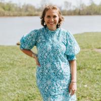 A photo of Anne Sydney smiling as she stands with one hand on her hip in front of the lake at the park.