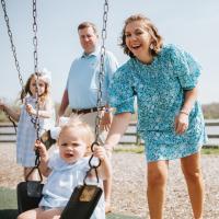 A photo of Anne Sydney and Jesse pushing their children on a swing set. Their oldest daughter, Winnie (left), is a young white girl with medium-length blonde hair. She is wearing a blue and white gingham dress that has a white collar, and a white bow in her hair.