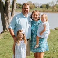 A photo of the Parrish family smiling as they pose together for the camera in front of a lake at the park.