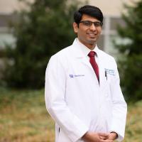 A photo of Dr. Chaitanya Iragavarapu smiling as he stands outside in front of a building on the UK HealthCare campus. He is a South Asian man with short black hair. He is wearing a white lab coat over a white button-up shirt with a red tie.