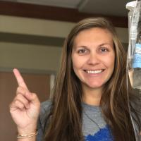 A photo of Anne Sydney smiling while posing with her IV bag and pointing her index finger in the air.